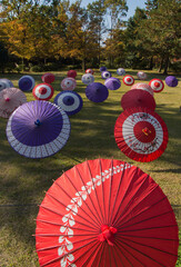 芝生の広場に色とりどりの和傘が沢山並べられ紅葉の木々が背景になっている風景