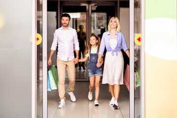 Fototapeta premium A family, consisting of a father, a mother, and a young daughter, walks out of a shopping mall, carrying shopping bags. The parents are holding hands with their daughter, and they are all smiling