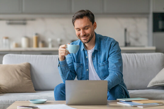 A man sits on a couch at home, working on a laptop while sipping a cup of coffee. He is wearing a casual blue shirt and smiling. He is working in a relaxed, comfortable setting.