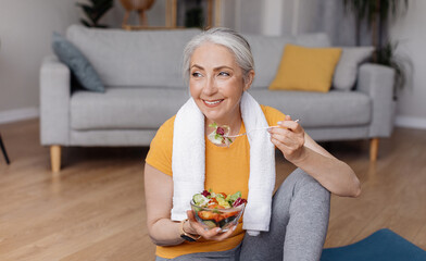 Healthy lifestyle concept. Happy senior woman eating fresh vegetable salad, sitting on yoga mat...