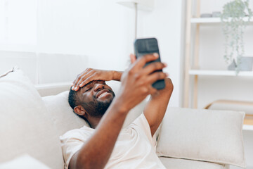 Happy African American man sitting on a black sofa, holding a mobile phone and typing a message The modern apartment provides a cozy background for his relaxed lifestyle and connection to the internet