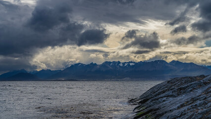  A picturesque snow-capped mountain range of the Andes against the sky and dark clouds. In the foreground is the water surface of the Beagle Channel, a rocky island. Argentina. Tierra del Fuego 