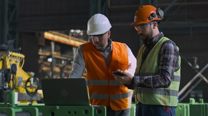 Two heavy industry engineers in uniform and helmets stand at manufacturing factory, work using laptop and mobile phone. Mature supervisor and adult employee have discussion at plant. Slow motion. - Powered by Adobe