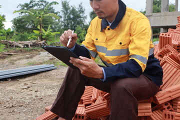A male engineer at a construction site wears a hardhat and reflective clothing, reviewing...