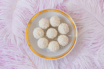 White Chocolate Candy With Coconut Topping On White Feather and pink background, sweet balls on plate flat lay. Tasty gift for any celebration and anniversary