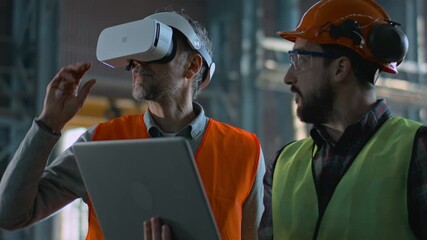 Two professional industry technicians wearing safety uniform and hardhats stand at manufacturing factory, use laptop and AR headset. Mature engineer and adult employee work in warehouse. Slow motion.