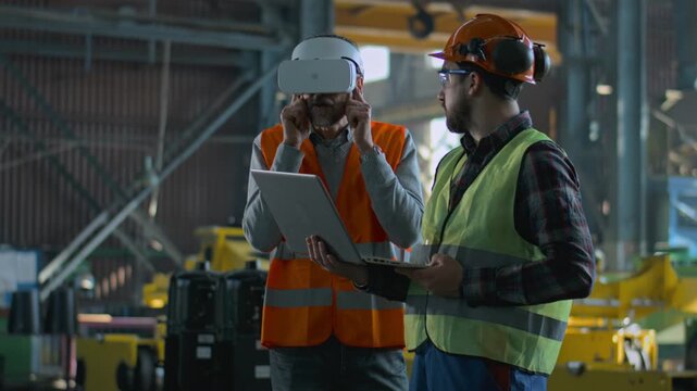 Two professional industry technicians wearing protective uniform and helmets stand at manufacturing factory. Mature engineer uses VR headset, adult employee work in laptop computer. Slow motion.
