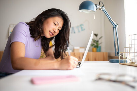 Asian female college student doing homework writing on paper at home. Chinise teen girl studying at home with laptop. - Powered by Adobe
