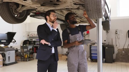 Mechanic in working overalls explaining car inspection details to business professional in auto repair shop. Businessman observing vehicle maintenance on lift with thoughtful expression.