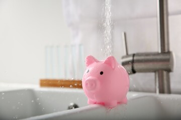 Water saving concept. Stream of water flowing from tap onto piggy bank in kitchen