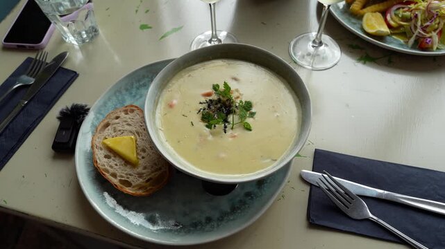 A bowl of seafood chowder served with a slice of bread and butter, accompanied by a glass of white wine and a plate of fish and chips.
