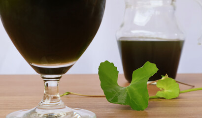 Centella asiatica fresh leaves and shoots sit on a table between a glass and a jar of dark green water. white background

