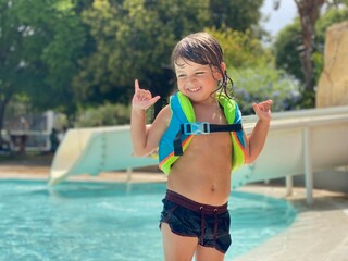 A laughing 3-year-old Caucasian boy wearing a life vest, standing by the pool with horizontal copy space.
