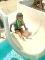  A smiling 3-year-old Caucasian boy enjoying a water slide on a sunny day at the pool.
