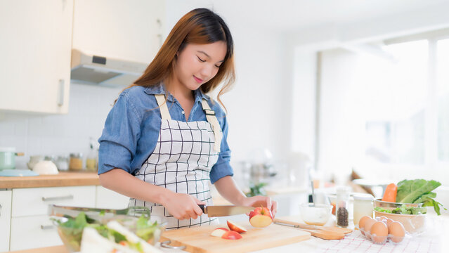 Young asian woman use knife to cutting slices apple on cutting board while preparing fresh vegetables salad in bowl and cooking healthy breakfast food in modern kitchen with healthy lifestyle at home