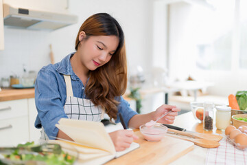 Young asian woman taking notes about food recipes in notebook and eating yogurt while preparing vegetables salad and cooking healthy breakfast food in modern kitchen with healthy lifestyle at home