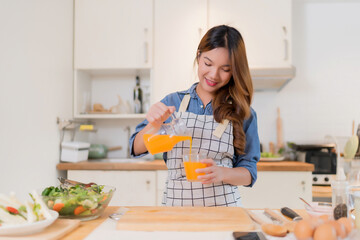 Young asian woman pouring orange juice from jug into the glass while preparing vegetables salad and cooking healthy breakfast food for homemade snack in modern kitchen with healthy lifestyle at home