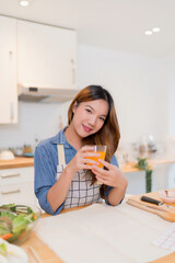 Young asian woman holding glass of orange juice and drinking fresh fruits while preparing vegetables salad and cooking healthy breakfast food in modern kitchen with healthy lifestyle at home