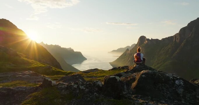 Woman overlook Senja fjord view from mountain in midnight sun glow; drone reveal