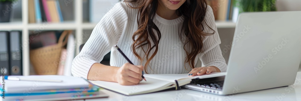 Poster Focused woman taking notes while studying at a desk in an office environment. She is using a laptop and has an open notebook, which creates a productive and professional atmosphere.