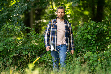 Fototapeta premium Confident lumberjack works in summer forest. Man with an ax. Woodcutter in plaid shirt in forest. Hipster with axe. Lumberjack with an axe. Bearded lumberjack. American lumberjack with hatchet.