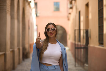 Young beautiful woman with brown hair, sunglasses and blue jacket suit, self-confident, thumbs up giving the ok. Concept beauty, fashion, trend.