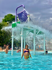 A 9-year-old Caucasian girl stands under a water bucket splash area, having fun at a pool on a sunny day.