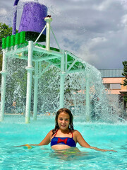 A close-up of a smiling 9-year-old Caucasian girl standing under a water bucket splash area, enjoying a sunny day at the pool.