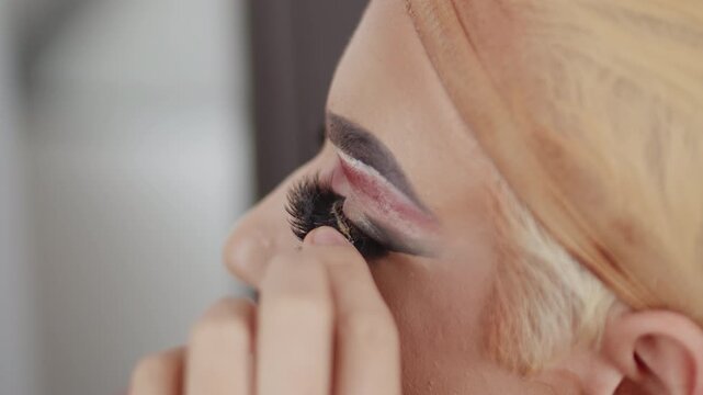 Young performer in drag queen makeup applying false eyelashes on his eyelids in the dressing room during preparation for the performance close up