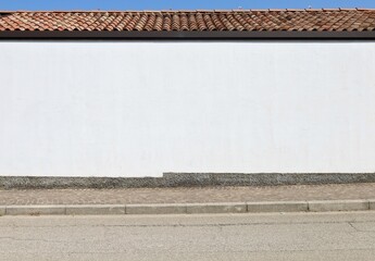 White plaster house facade at the roadside with rooftiles on top. Sky above, sidewalk and street in...