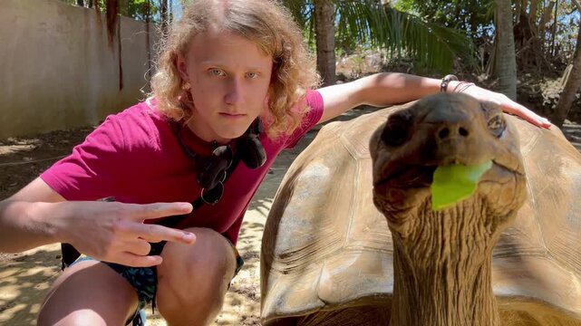 Tourist boy posing for photo with Aldabra giant tortoise endemic species - one of the largest tortoises in the world in the zoo nature park on Mauritius island. Then both noticed it was video shooting