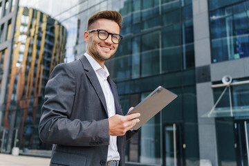 A smiling young businessman in a gray suit uses a tablet outdoors, standing in front of modern office buildings.