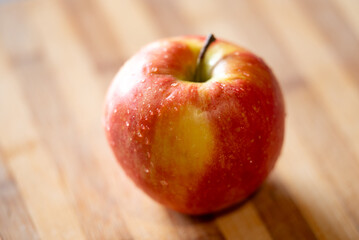 A red apple with yellow spots laying on a wooden table, close up. One apple with water drops on it, lightened by sunlight, concept of healthy fruit
