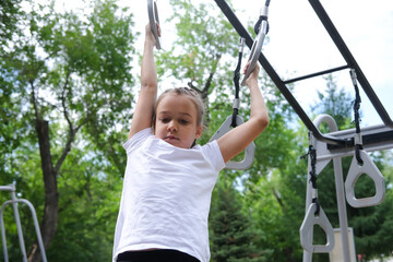 Fototapeta premium Girl performs exercises on gymnastic rings on an outdoor sports ground on sunny day. Child is training outdoors. Concept of children's healthy lifestyle, children's leisure time on vacation.
