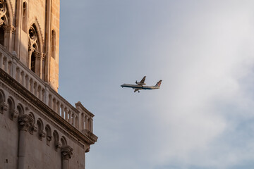 Commercial plane flying low over historic bell tower of Cathedral of St. Lawrence (San Lorenzo) against dramatic sky in old town Trogir, Split-Dalmatia, Croatia, Europe. Tourist travel destination