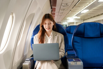 An Asian businesswoman travels abroad by plane, sitting near the window. She reads a book and uses her computer, utilizing her airline credit card to manage travel expenses efficiently.