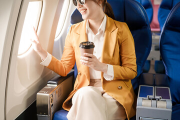 An Asian businesswoman travels abroad by plane, sitting near the window. She reads a book and uses her computer, utilizing her airline credit card to manage travel expenses efficiently.