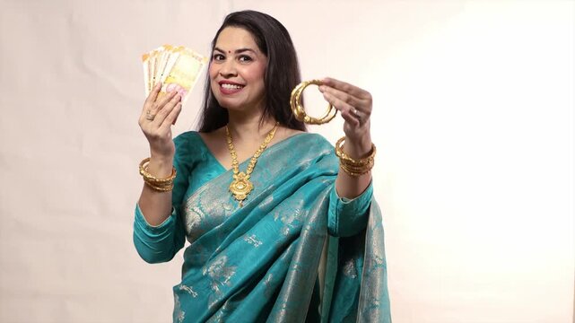 Happy young Indian woman wearing saree holding gold jewelry bangle ornaments in hand and rupee notes standing against white background, Concept of Gold Loan.Investment in asset class