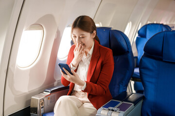 An Asian businesswoman travels abroad by plane, sitting near the window. She reads a book and uses her computer, utilizing her airline credit card to manage travel expenses efficiently.