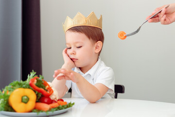 Cute little boy siting at the table and refusing to eat vegetables at home