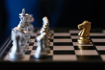 Golden knight placed against many silver chess pieces on chessboard with businessman in background