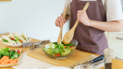 Asian women using wooden spoon to mixing ingredients and slices of fresh vegetable in the glass bowl to cooking breakfast meal and vegetable salad while making homemade healthy food lifestyle in home