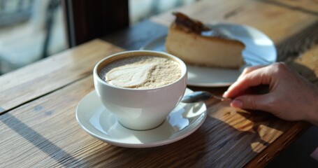 Woman stirs cappuccino with milk froth leaf during breakfast in coffeeshop closeup. Lady with coffee cup and cheesecake at wooden counter. Cappuccino drink