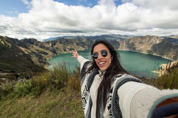 Naklejka premium Latina young traveler taking a selfie with a camera in front of Quilotoa Lagoon in Ecuadorian Andes