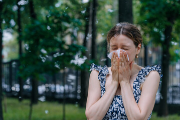 Woman sneezes into a tissue while standing outside in a park, surrounded by airborne allergens like...