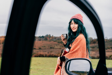 Hipster Woman sits on car's hood, wrapped in warm Shawl Wraps Poncho Cape and hat and enjoying warm drink, while overlooking rural landscape during road trip. Cold season travel, vacation concept