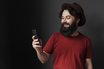 A cheerful bearded man sporting a stylish hat looks at his phone screen with a smile. Wearing a red t-shirt, simplistic background, epitomising casual connectivity and modern lifestyle.