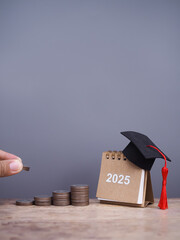 Close up hand putting coin into stack of coins and 2025 Desk calendar with graduation hat. The concept of saving money for education, student loan, scholarship, tuition fees in year 2025