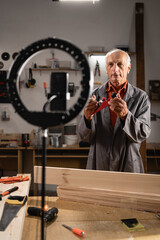 Elderly carpenter with phone on tripod recording vlog, communicating with followers, livestreaming...