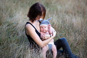 Mother hugging newborn baby boy in her hands outdoors in summer in sun rays, sitting in grass